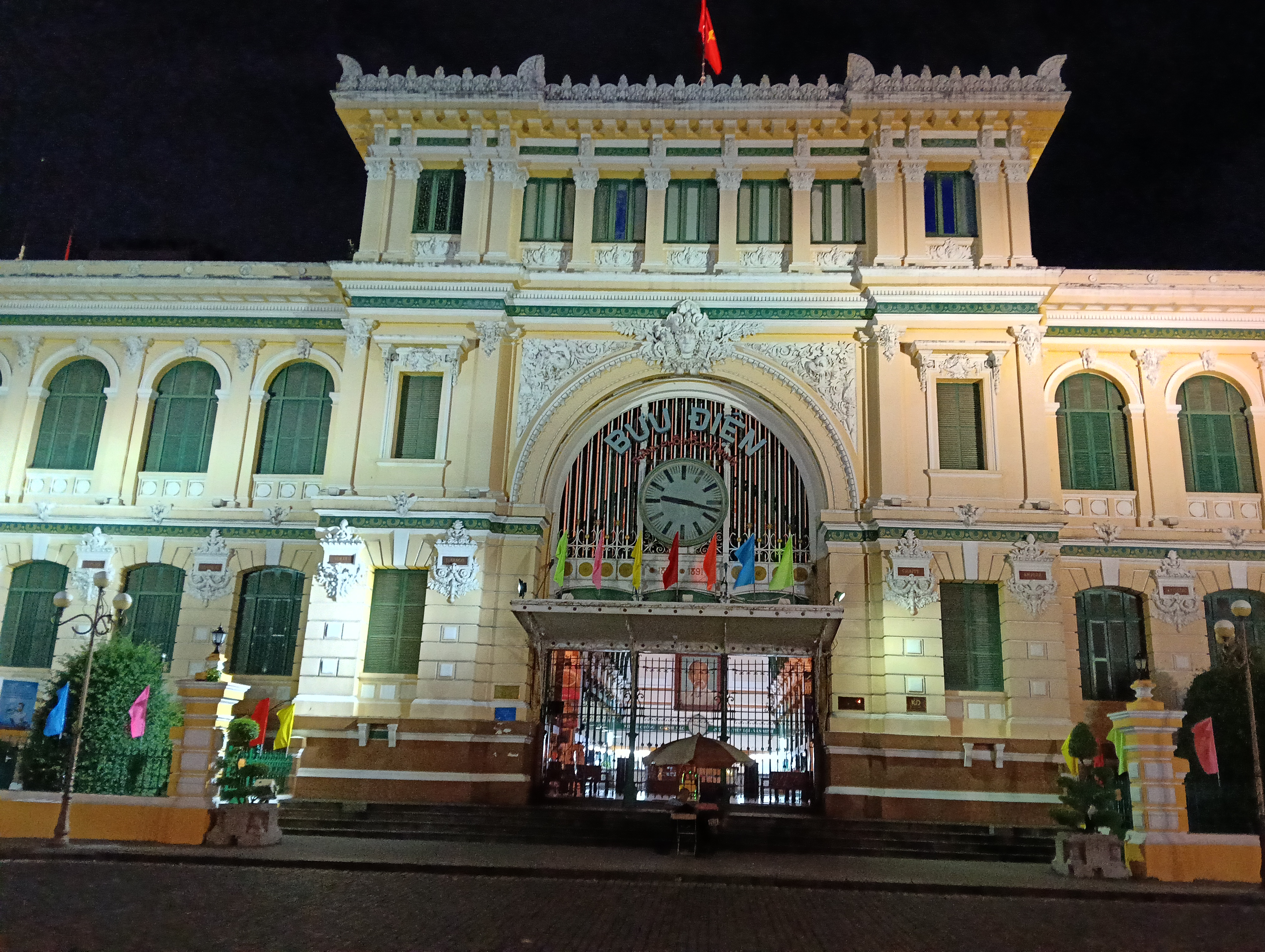 胡志明市中央郵政夜景，黃色法殖民時期建築正面打燈，正立面大鐘清晰可見、上方掛越南國旗