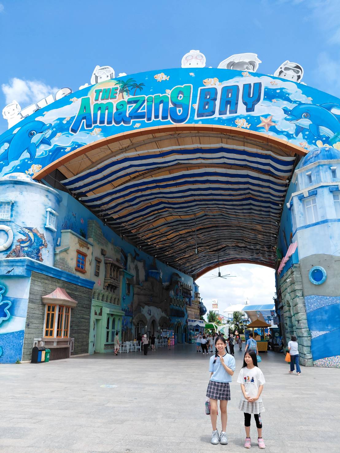 The Amazing Bay 神奇海灣水樂園主入口大門，藍色海洋主題拱門上寫著大大的 The Amazing Bay 字樣，兩個女兒站在前面合照